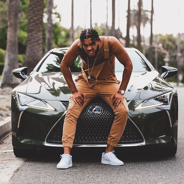 Sarunas J. Jackson in a brown dungaree and white sneakers posing in front of his luxurious car.